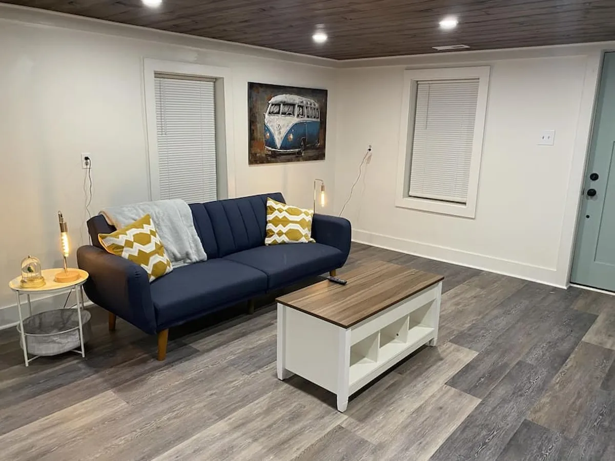 A stylish living room featuring a blue sofa with a yellow patterned pillow, a wooden coffee table, and framed artwork on the wall. The space feels modern and comfortable, with ample seating for relaxation or socializing and large windows providing abundant natural light.
