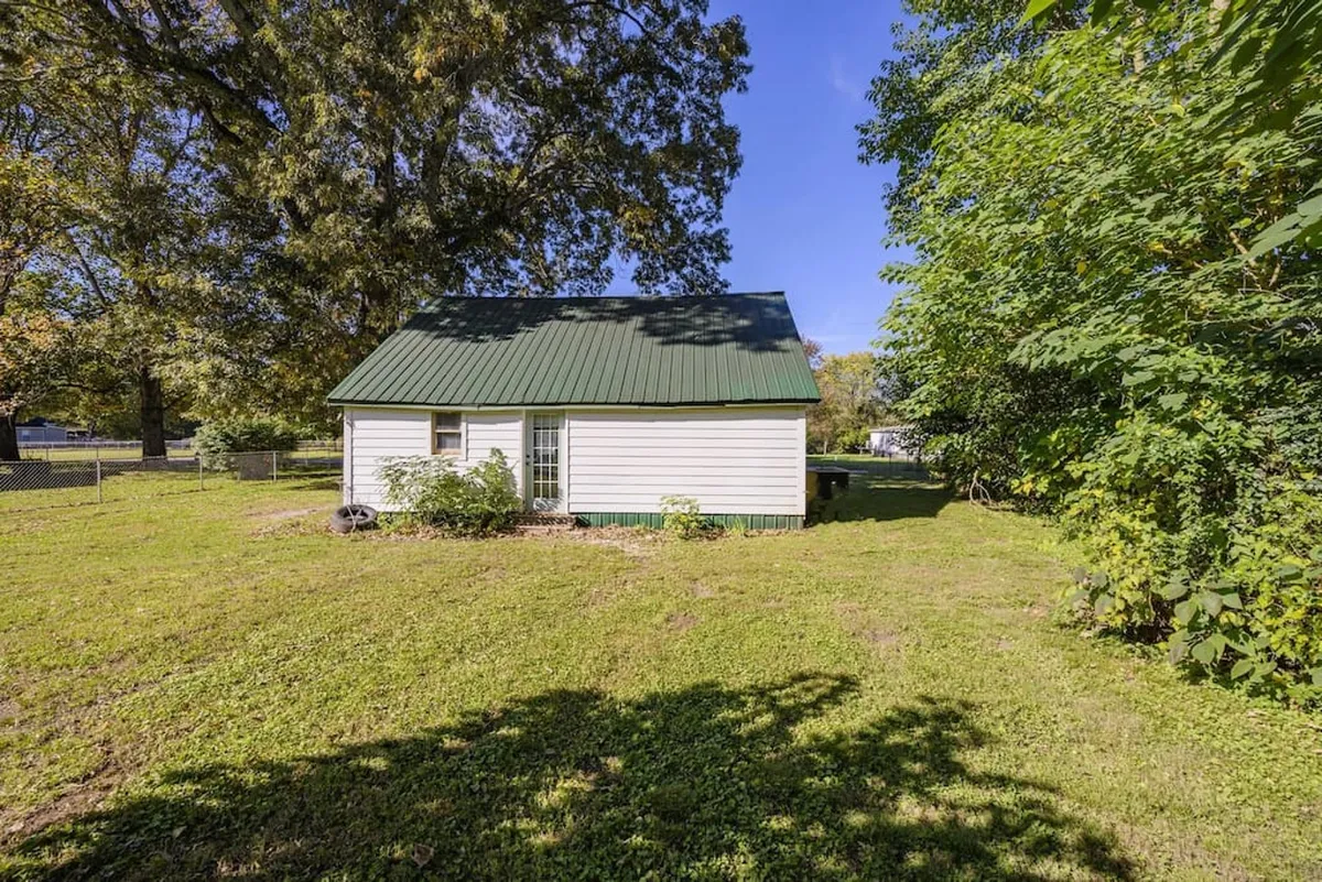 This image showcases a small house with a green roof nestled among mature trees and surrounded by an expansive open yard. The open grass provides plenty of space for children to play, and the setting is clearly designed for outdoor gatherings and relaxing in nature.