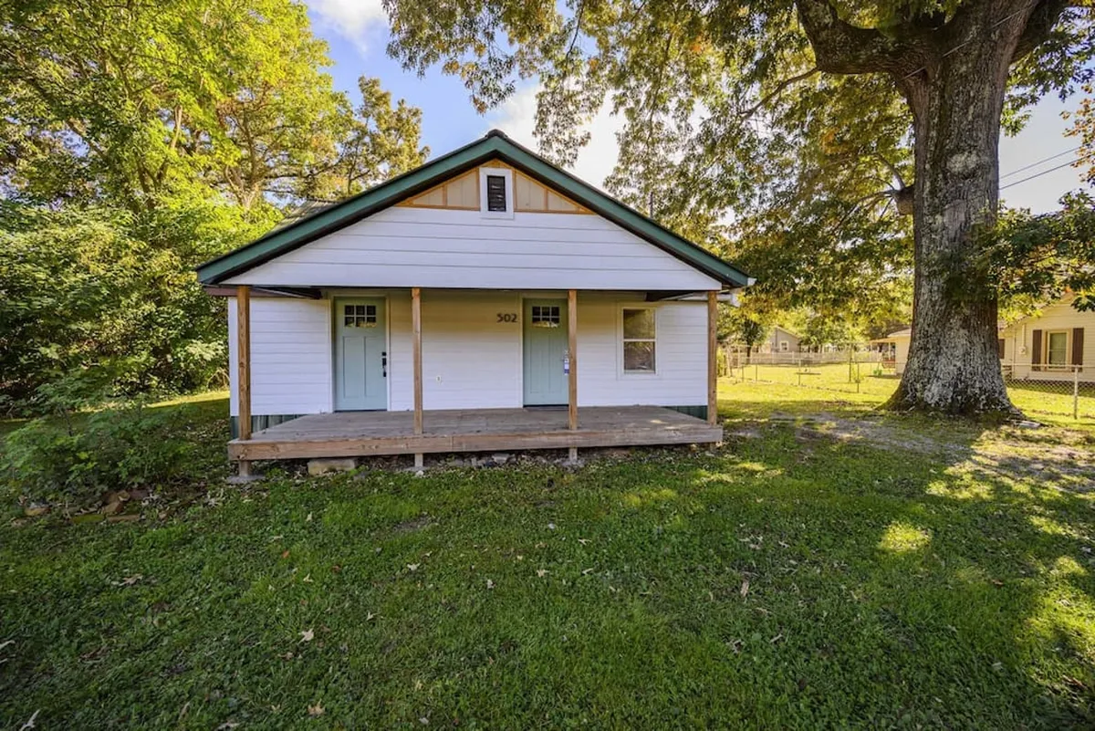The image showcases another angle of the front porch and entryway, complete with a charming green door, lush grass, and the large tree offering shade. The setting is perfect for social gatherings and enjoying quiet mornings before venturing out.