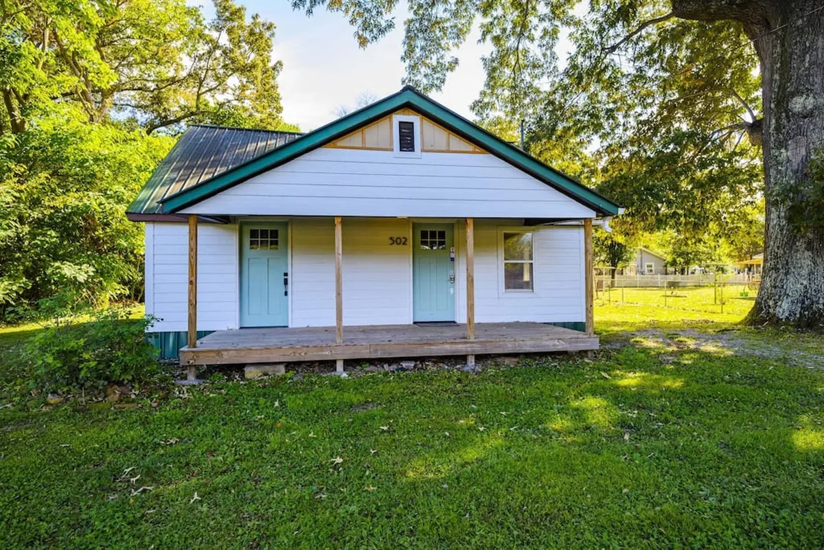 A charming exterior view of the home, featuring a front porch with blue doors, a green roof, and inviting outdoor seating. The porch overlooks a yard, suggesting a seamless transition between indoor comfort and outdoor enjoyment.
