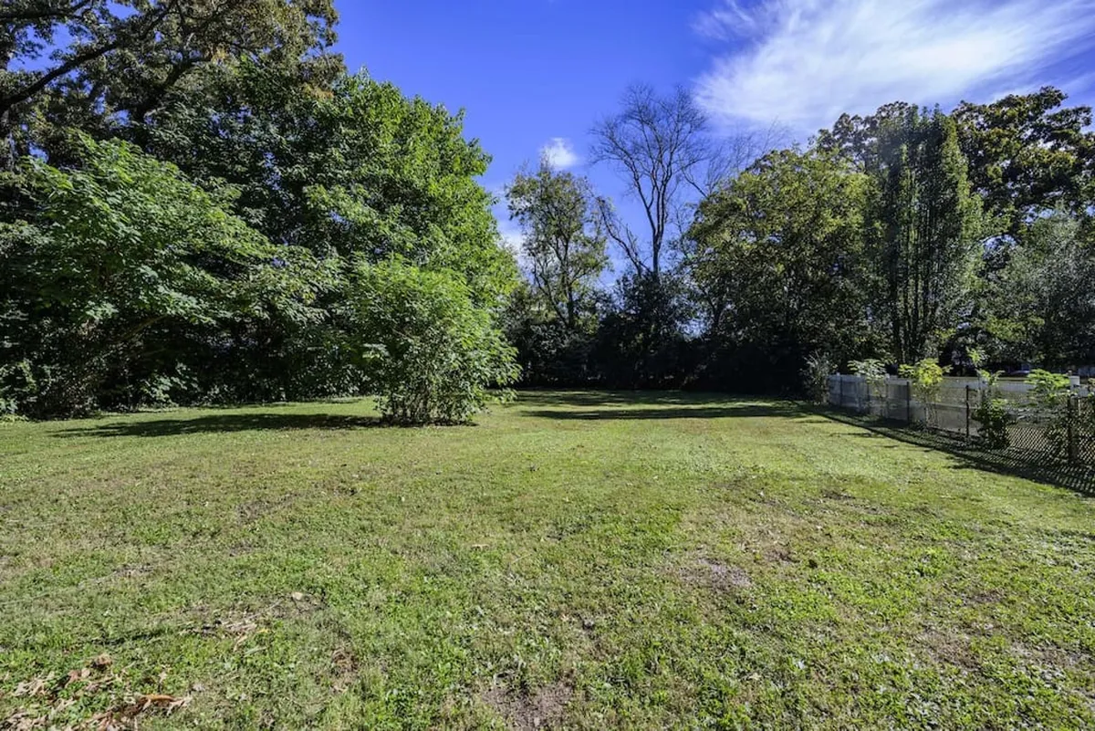 A tranquil, open grassy area bordered by lush greenery and trees, perfect for picnicking, playing, or quiet reflection. The setting feels welcoming and safe for families or groups to enjoy the outdoors.