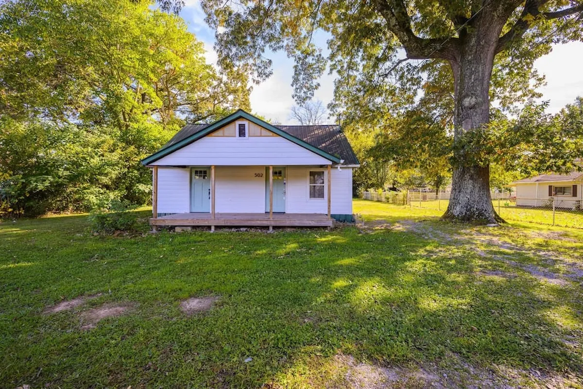 This image captures the welcoming front porch of the home, shaded by a large tree and set against a green lawn. The scene invites relaxation outdoors, offering a glimpse of where guests might unwind after an evening out or gather before heading to a concert or local event.