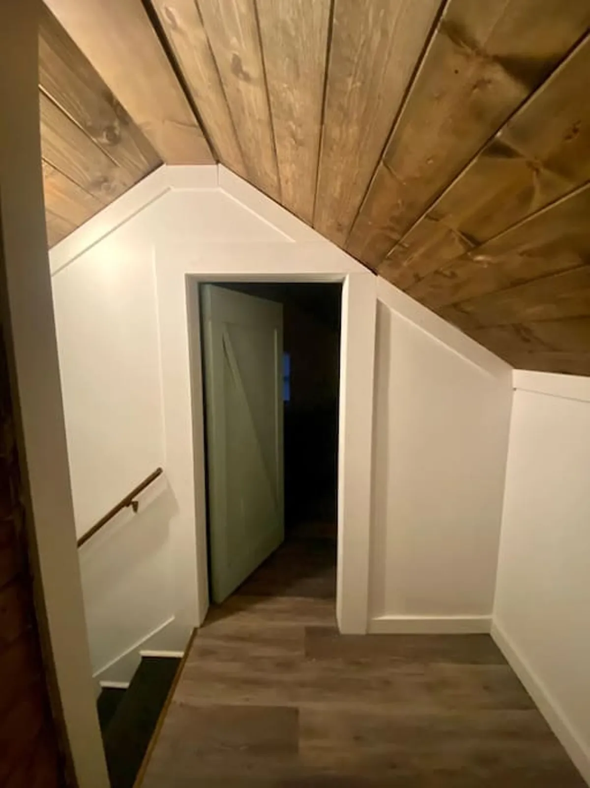 This image features a staircase leading to an upper floor, visible through a doorway beneath a warm wooden ceiling. The scene gives a sense of movement and discovery, with someone walking towards the next room. The lighting is inviting, evoking a sense of anticipation and exploration within the home.