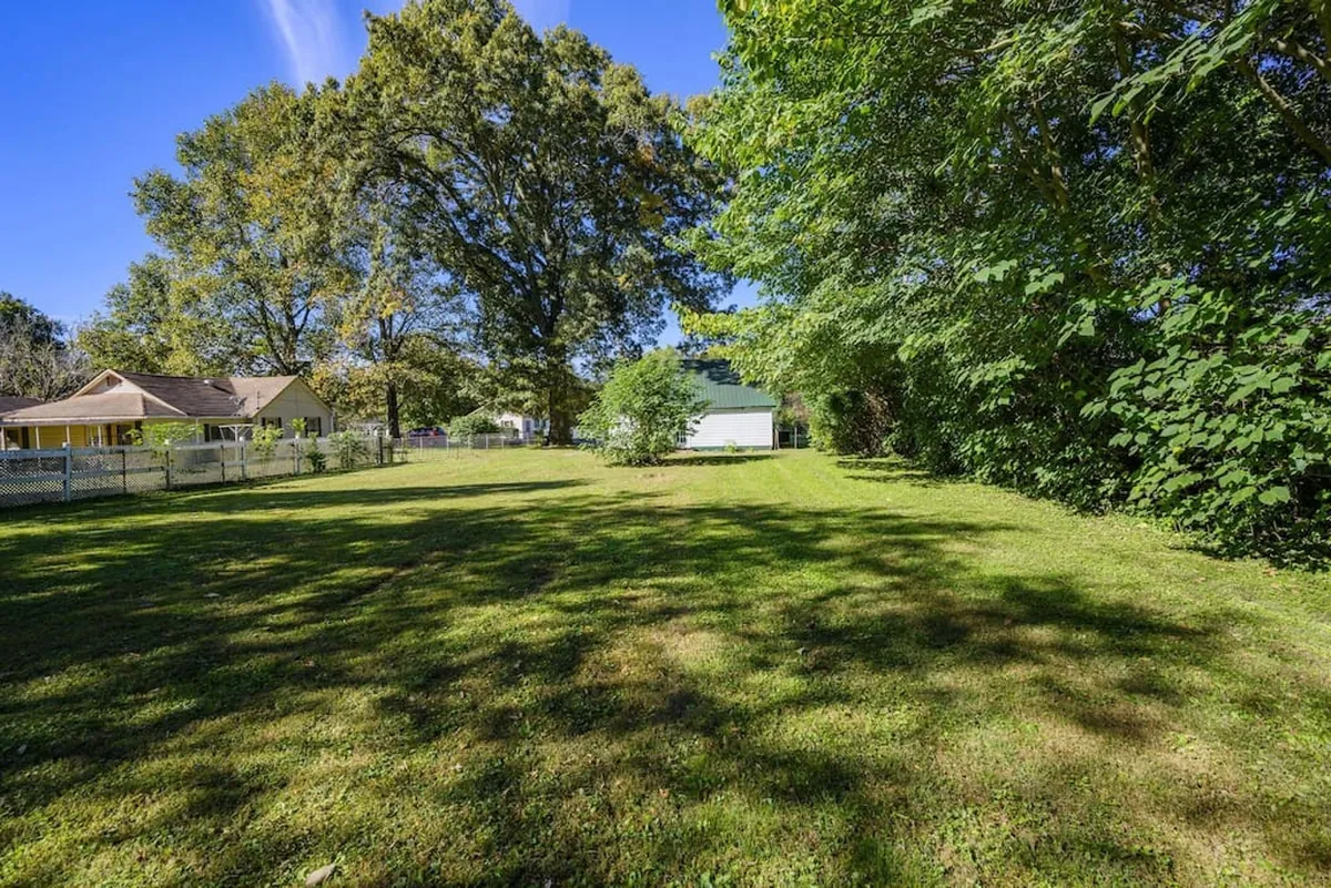 A lush green lawn surrounded by large trees and distant structures, offering a spacious, family-friendly yard setting. The scene suggests a welcoming space ideal for outdoor games, gatherings, and playtime for children.