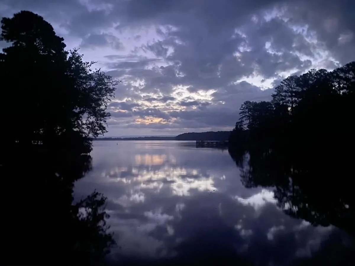 A tranquil view over a calm water surface at dusk, with a dramatic sky and silhouetted trees lining the horizon. The scene evokes a peaceful, natural setting perfect for quiet walks or romantic moments by the water.