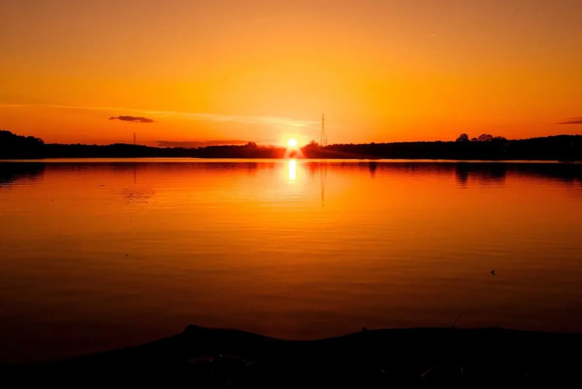A breathtaking sunset reflected on the water, with distant hills and a silhouetted landscape creating a serene and romantic mood. The scene invites relaxation and appreciation of natural beauty.